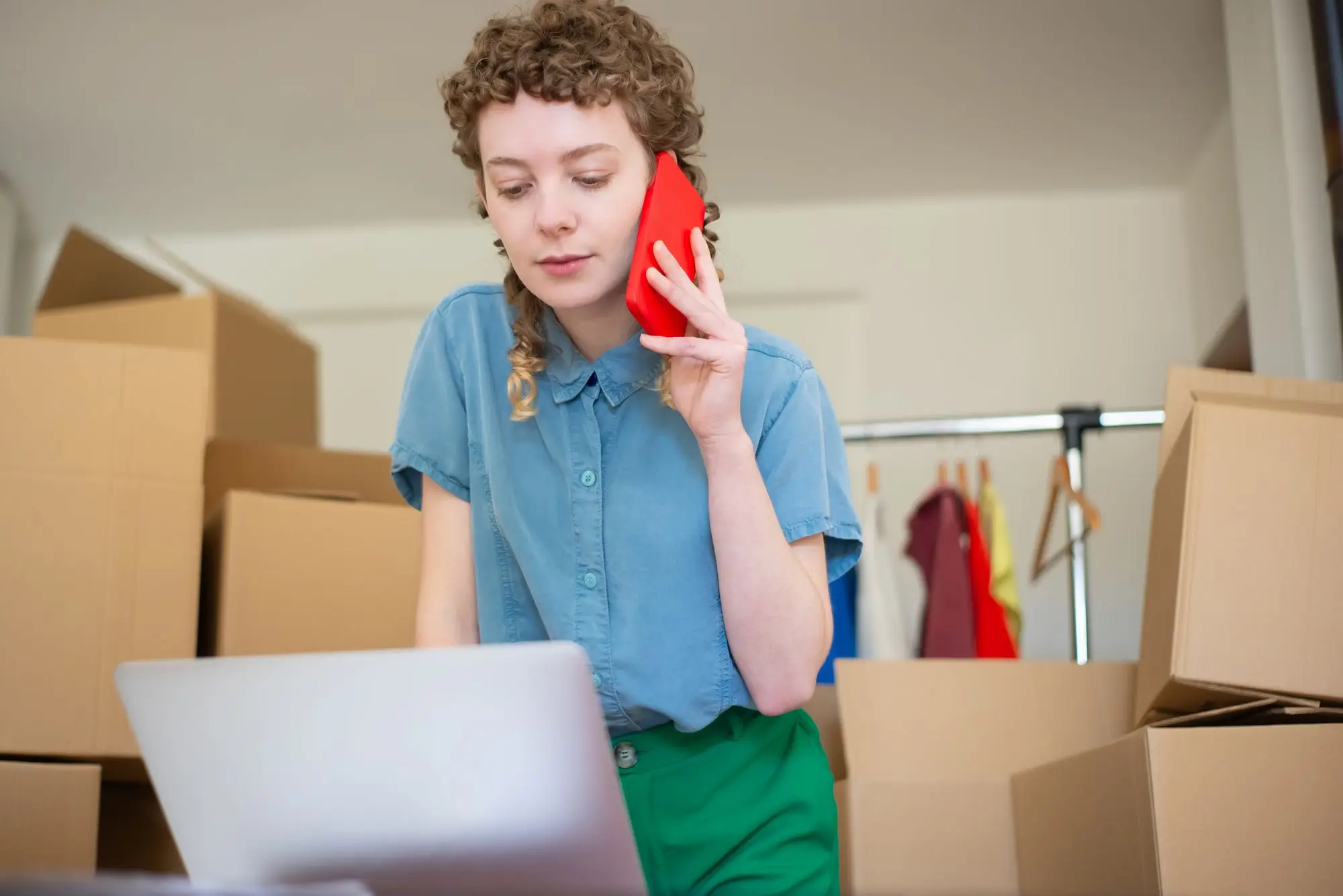 Online clothing store owner on a phone call while checking her laptop among stock boxes