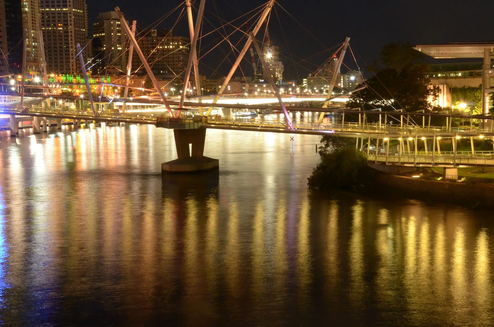 Kurilpa Bridge lit up over the Brisbane River at night, with city buildings behind